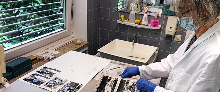 Eine Frau in Arbeitskleidung steht an einem Tisch, auf dem Fotografien liegen.