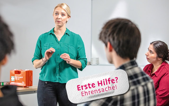 Eine blonde Frau in türkisem Oberteil steht in einem Seminarraum und redet. Um sie herum sitzen Personen, die ihr zuhören. Neben der Frau steht ein Erste-Hilfe-Koffer.