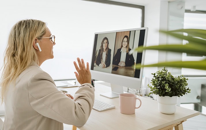 Eine blonde Frau mit Brille von schräg hinten, die an einem Schreibtisch sitzt und in Richtung des Bildschirms vor sich winkt, auf dem eine Konferenzschaltung mit zwei anderen Frauen zu sehen ist.