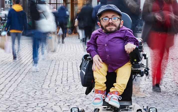 Der Aktivist und Buchautor Raúl Krauthausen sitzt in seinem Rollstuhl in einer Fußgängerzone. Er trägt eine gelbe Hose, ein lilanes Hemd, eine graue Kappe und eine Brille.
