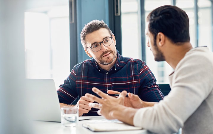 Zwei männliche Personen sitzen sich gegenüber und sprechen miteinander. Die eine Person sitzt mit dem Rücken zur Kamera und gestikuliert, während die andere gespannt zuhört.
