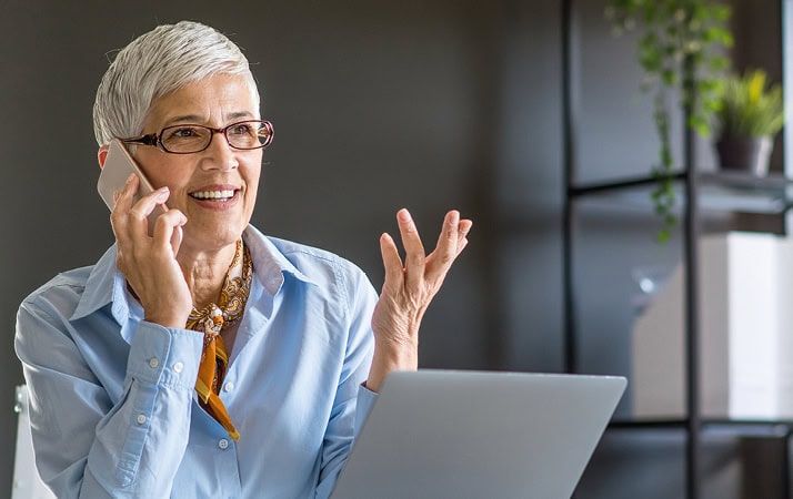 Eine Frau mit kurzen weißen Haaren und Brille hält sich ein Smartphone an ihr Ohr. Dabei lächelt sie und gestikuliert mit der linken Hand.