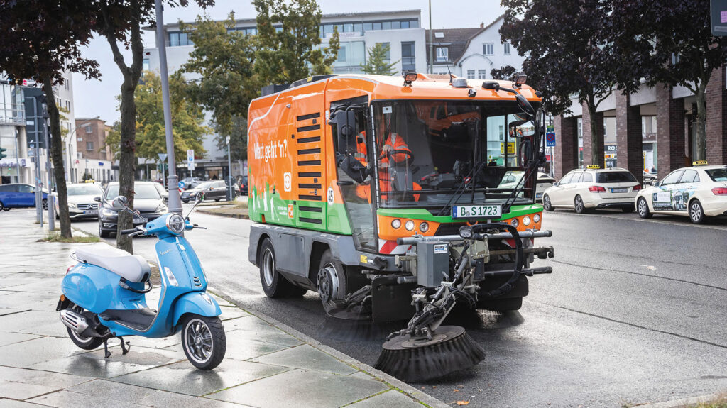 Eine orange Kehrmaschine reinigt die Fahrbahn entlang des Gehsteigs.