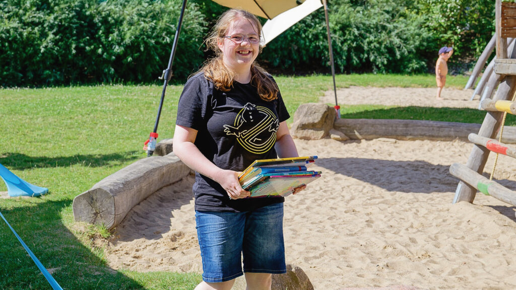 Laura Reyer geht mit einem Bücherstapel in der Hand durch den Garten der Kita. Sie trägt sommerliche Kleidung und hat eine Brille auf.