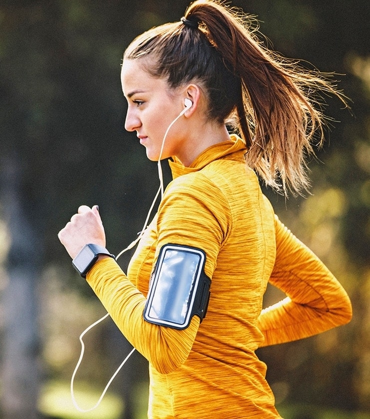 Eine junge Joggerin in gelber Jacke läuft draußen in der Natur. Sie trägt eine Smartwatch und ein Armband für ihr Handy.