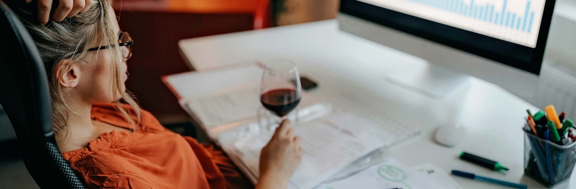 Eine Frau sitzt an einem Schreibtisch und schaut auf ein Diagram auf einem PC. Die hält ein Glas Rotwein in der Hand.