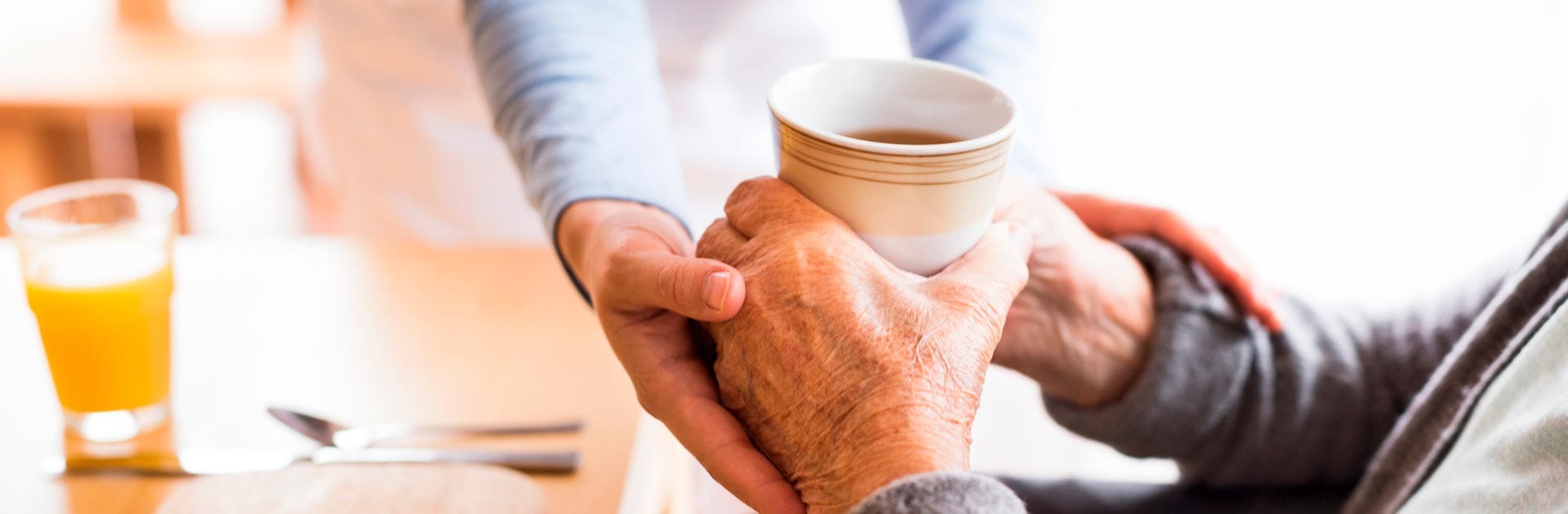 Eine Seniorin hält in den Händen eine Tasse Kaffee und wird dabei von einer Pflegerin unterstützt.