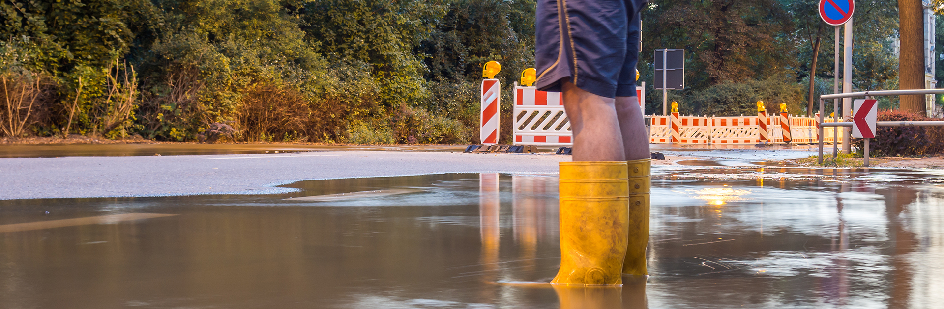 Eine Person mit kurzer Hose und gelben Gummistiefeln steht auf auf einer überfluteten Kreuzung