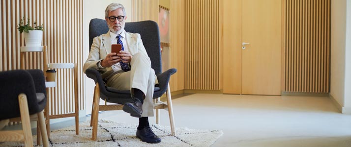 Ein Mann sitzt in einem dunkelgrauen Sessel im Flur eines Bürogebäudes. Die Wände sind mit Holz verkleidet, eine Tür ist ebenfalls aus hellem Holz. Der Mann trägt einen hellen Anzug, eine Brille und schaut gerade auf sein Smartphone.