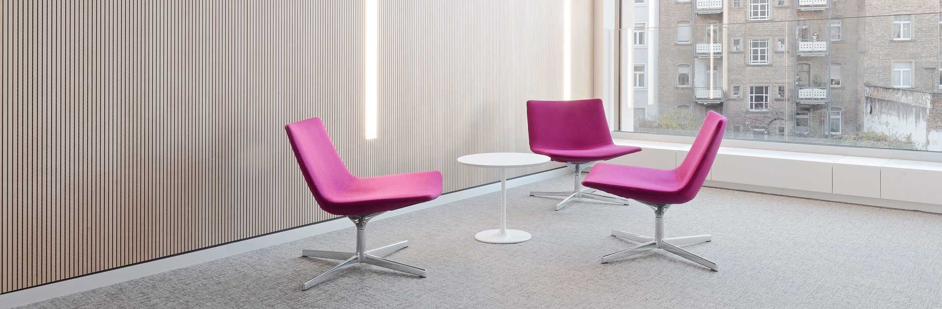 Drei violett gepolsterte Stühle sind um einen weißen, runden Tisch gruppiert. Im Hintergrund befindet sich ein bodentiefes Fenster. Links ist die Wand mit hellem Holz verkleidet. Der Teppichboden und die Decke sind hellgrau.