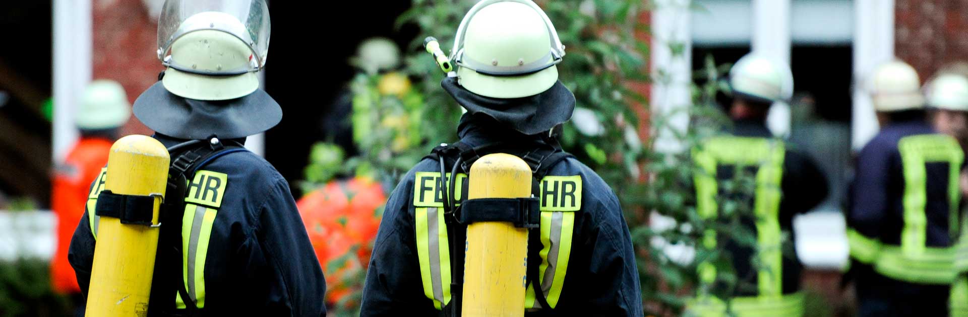 Man sieht zwei Feuerwehrmänner von hinten. Sie tragen volle Schutzkleidung: Weiße Helme, schwarze Schutzkleidung mit neon Streifen und gelben Sauerstoffflaschen.