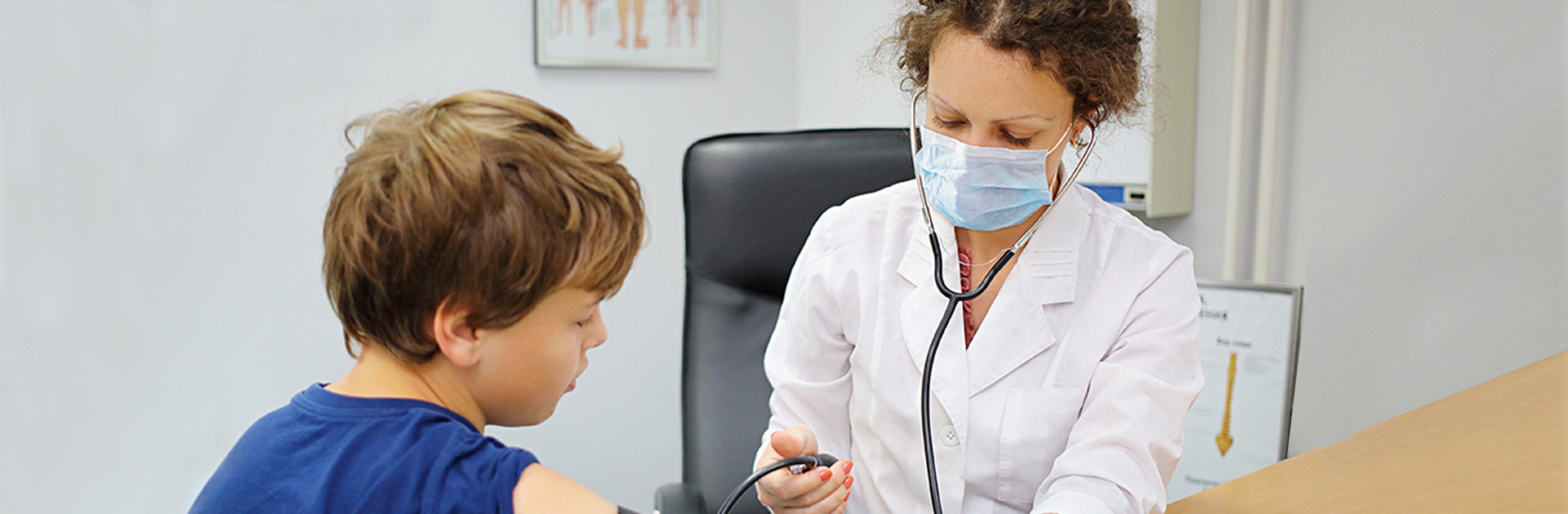 In einer Praxis misst eine Ärztin einem Jungen den Blutdruck. Sie trägt einen weißen Kittel sowie einen Mund-Nasen-Schutz. Der Junge trägt ein blaues T-Shirt und am Oberarm die Manschette des Messgeräts.