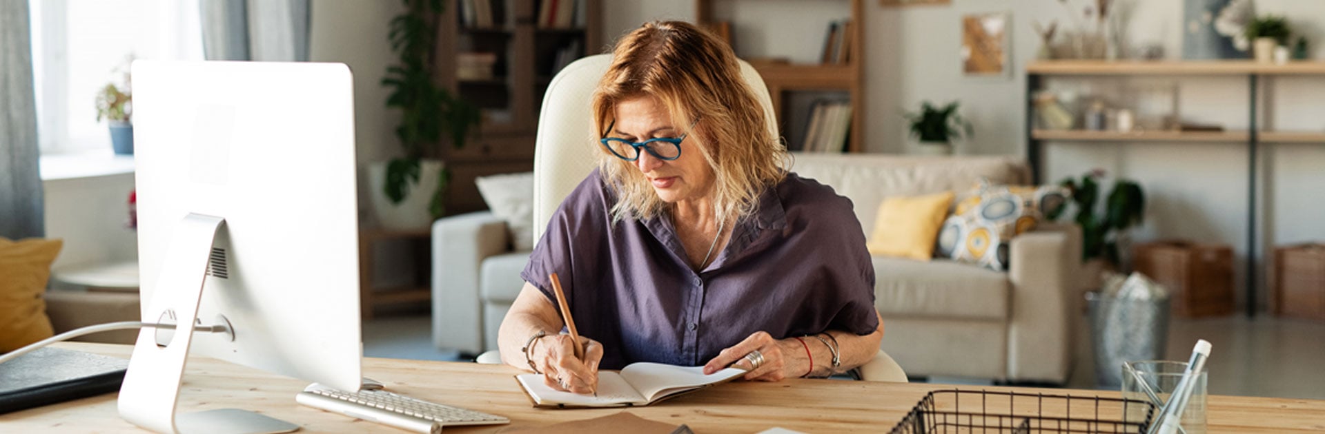 Eine Frau mit dunkelblondem Haar, und einer dunkelgrüner, dicker Brille schreibt am Schreibtisch in ein Buch. Sie sitzt in einem Raum, der sehr wohnlich eingerichtet ist.