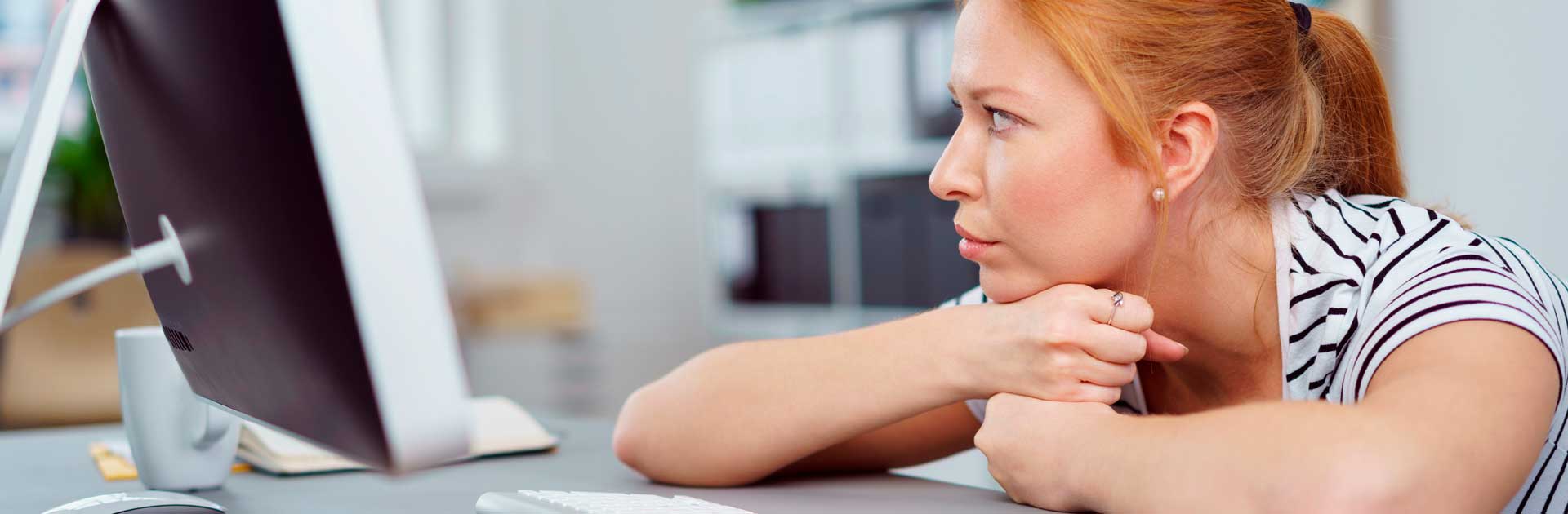 Eine junge Frau sitzt an einem Schreibtisch und blickt nachdenklich zur Seite. Das Kinn hat sie auf ihre Hände gestützt. Sie trägt ein schwarz-weiß gestreiftes T-Shirt und hat ihre roten Haare zu einem Zopf gebundenen. Auf dem Schreibtisch befinden sich eine Brille und eine Computertastatur.