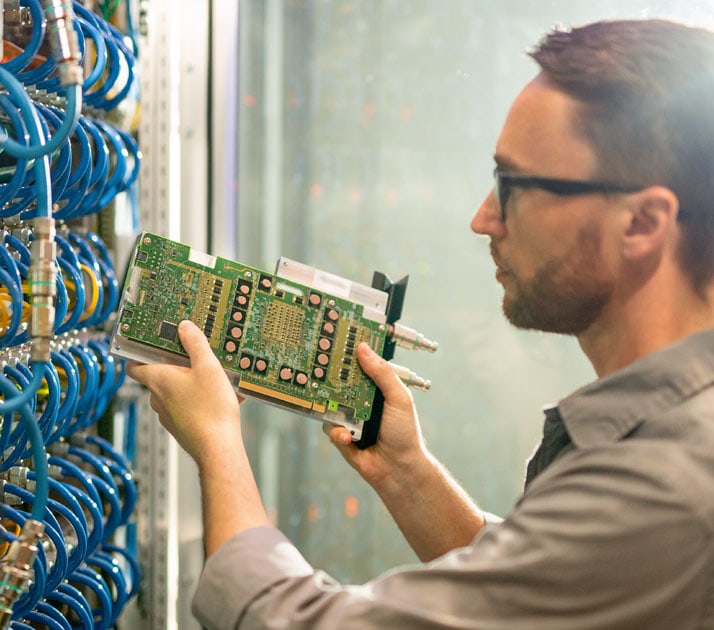 Ein IT-Experte steht vor einem Serverschrank, aus dem viele blaue Kabel herausschauen. Er steckt eine grüne Platine ein.