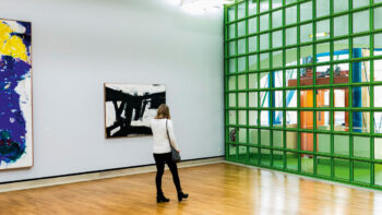 Eine Frau läuft durch den Ausstellungsraum der Staatsgalerie Stuttgart. Vor ihr hängen zwei großformatige Gemälde. Der Raum wird vor allem durch ein großes gläsernes Oberlicht beleuchtet.