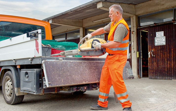 Ein Arbeiter in orangener Warnkleidung hebt ein Arbeitsgerät von der Ladefläche eines Fahrzeugs. Er befindet sich auf einem Betriebshof.