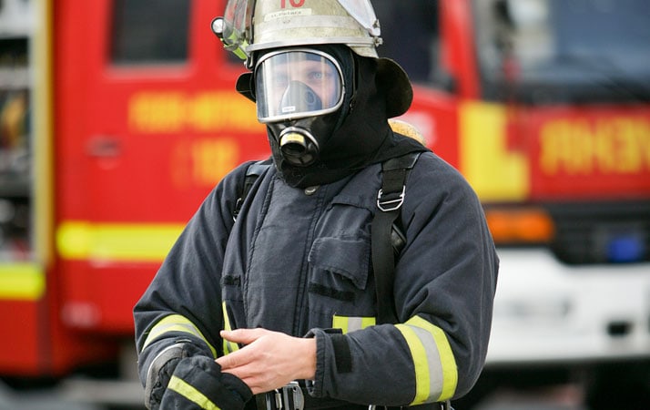Ein Feuerwehrmann in schwarzer Arbeitskleidung steht vor einem Feuerwehrauto. Er trägt Schutzhelm, ein Atemschutzgerät und den Gurt einer Absturzsicherung. Der Mann zieht gerade seine Handschuhe an.